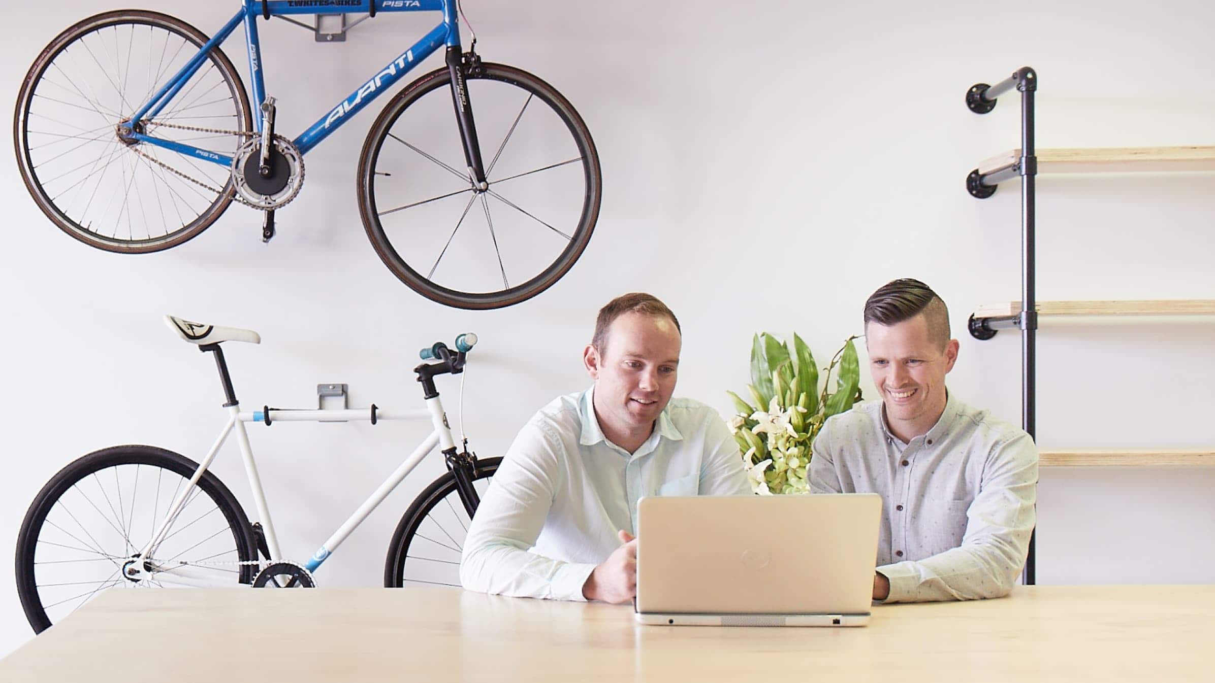 Peter Harris and Nathan Maisey sit working at a laptop, with two bikes and a set of shelves on the wall behind them.