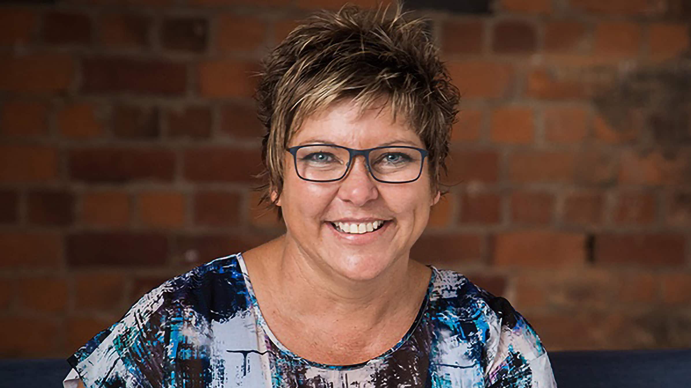 Di Crawford-Errington, owner of Ontrack Bookkeeping, smiling to camera against brick wall backdrop.