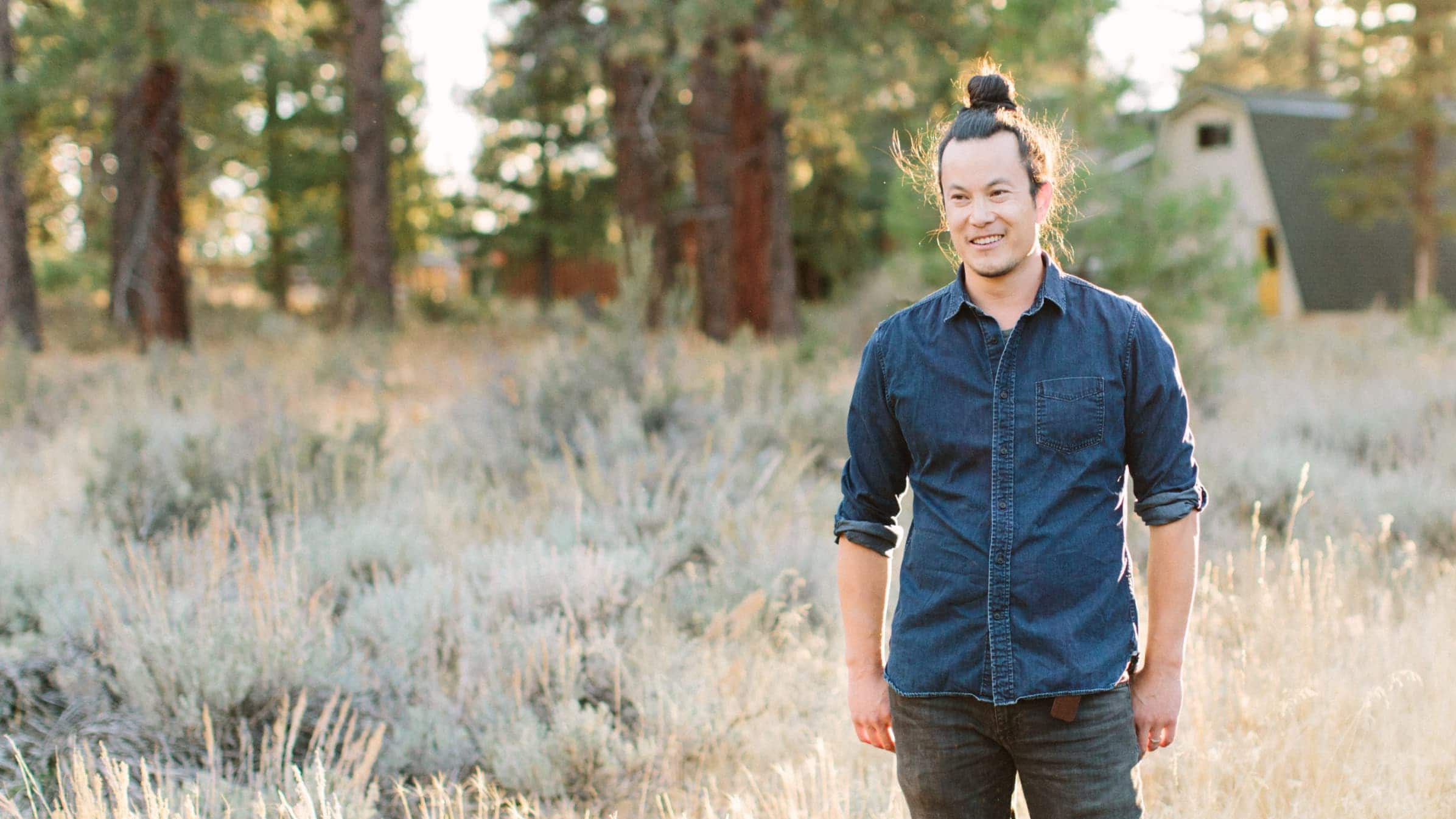 Burton Li, bookkeeper from Sutro Li, stands in a sunny field with trees and his home in the background.