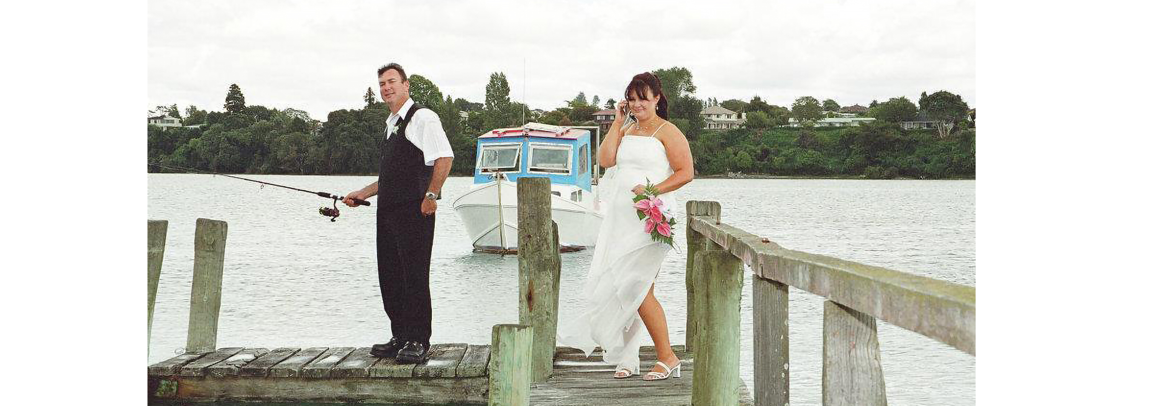 Janine Gartner standing on a pier in her wedding dress, with husband fishing beside her.