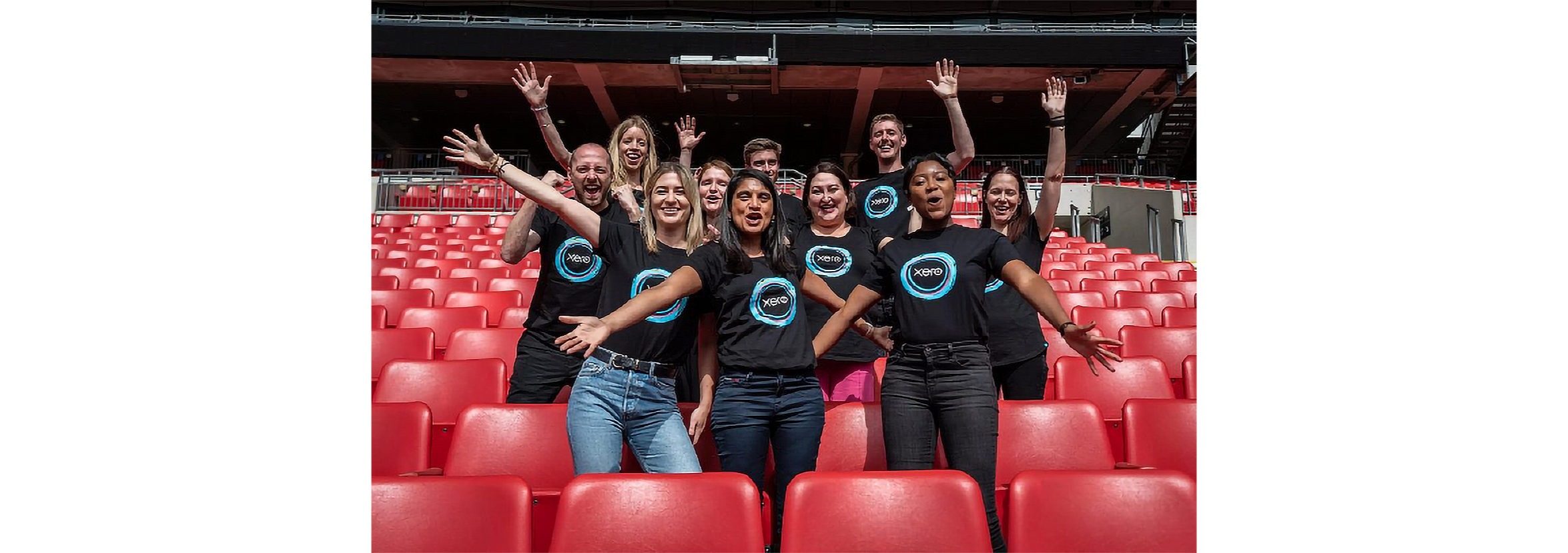 A group of employees in Xero t-shirts pose for the camera, with arms outstretched, in a large events arena.