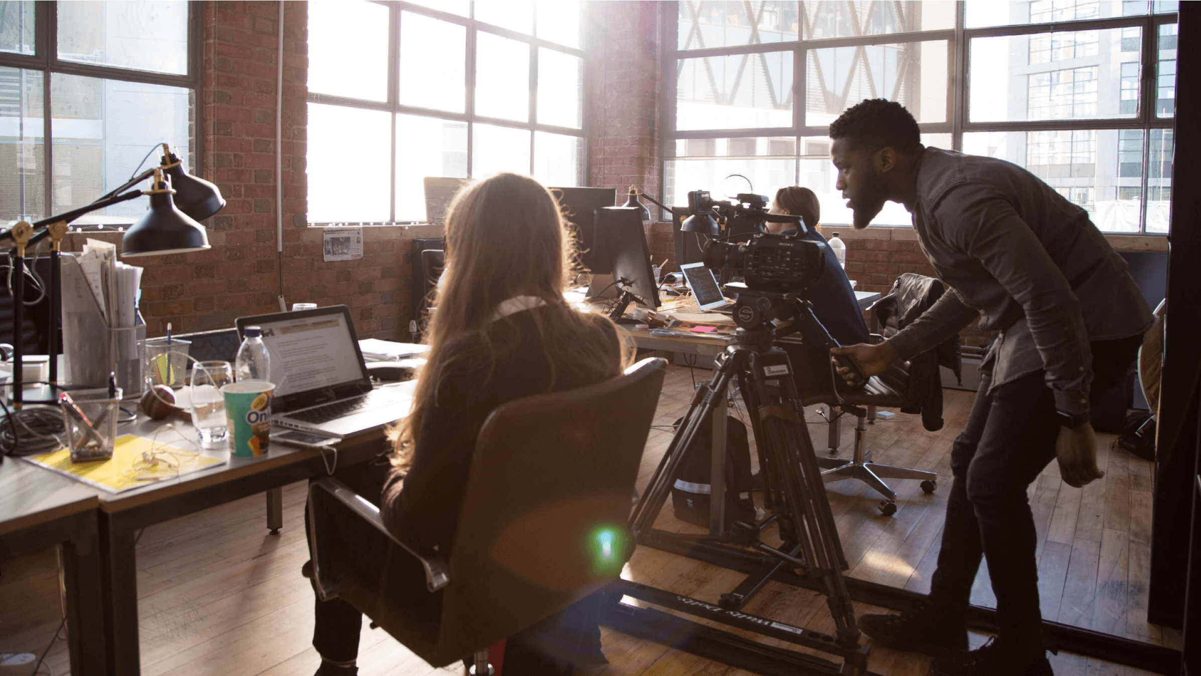 A small business owner sits in front of their laptop as a camera person records the story of their success.