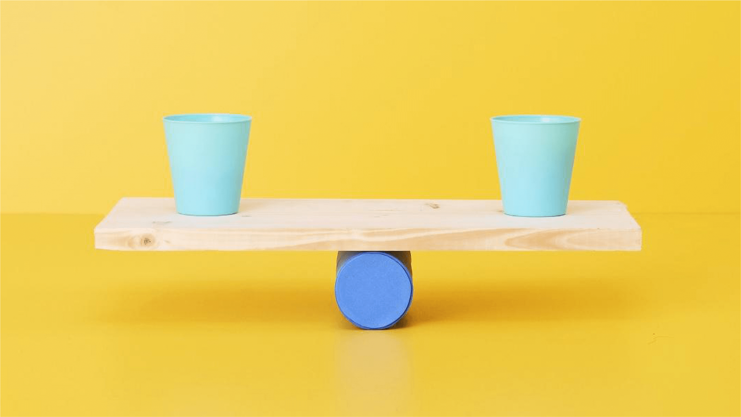 Two green cups sit on either side of a wooden plank balanced on top of a cylindrical roller.