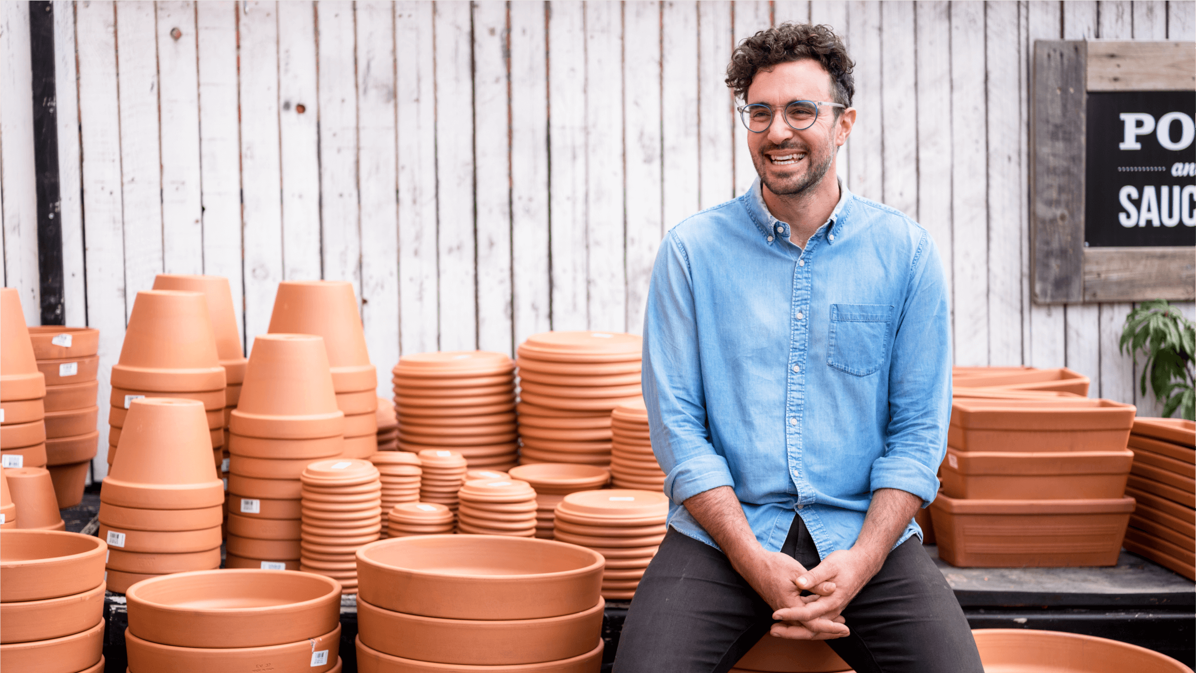 A business owner sits among various plant pots, smiling.