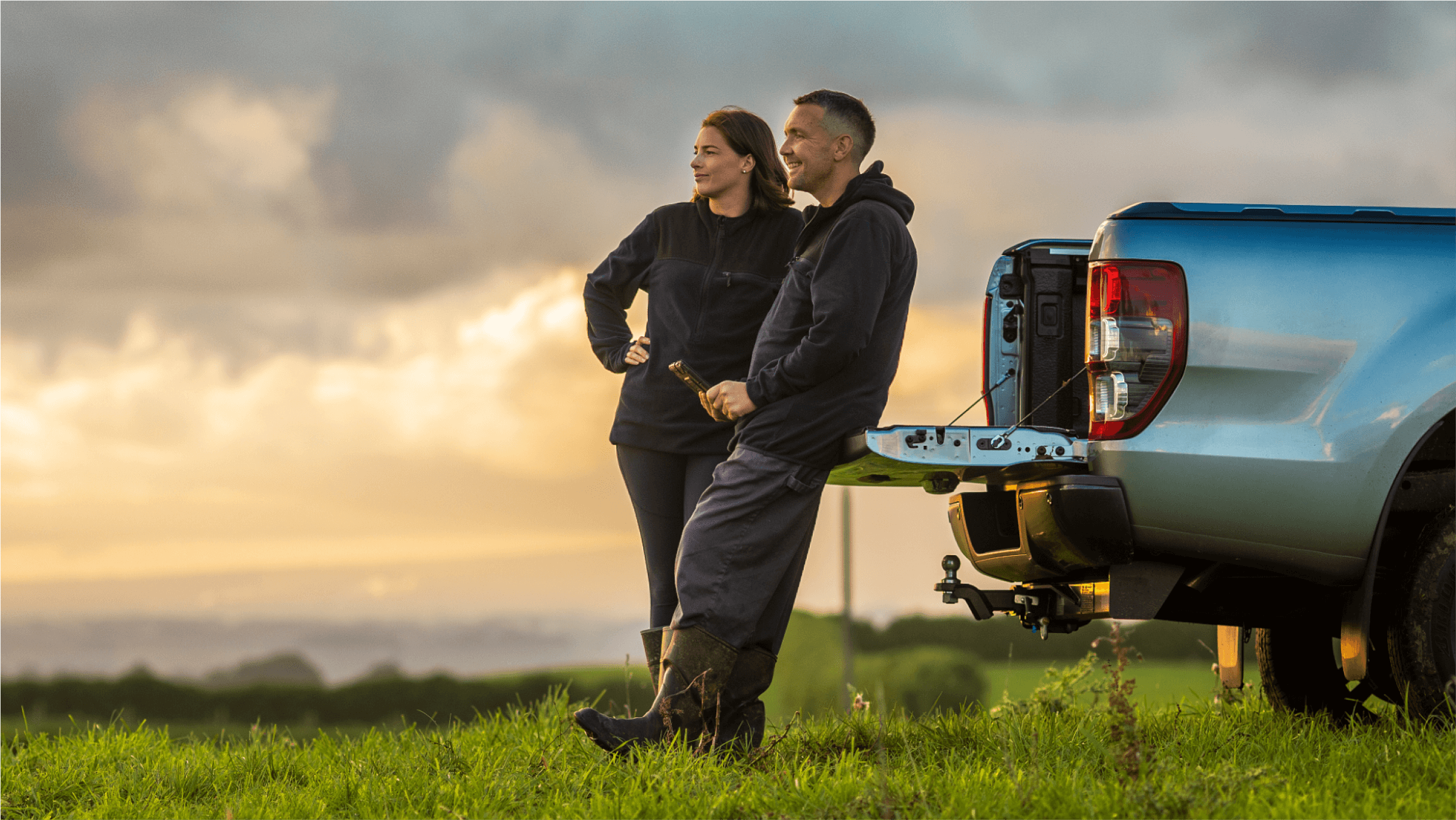 Two farmers lean on the open tray of a ute in a field.