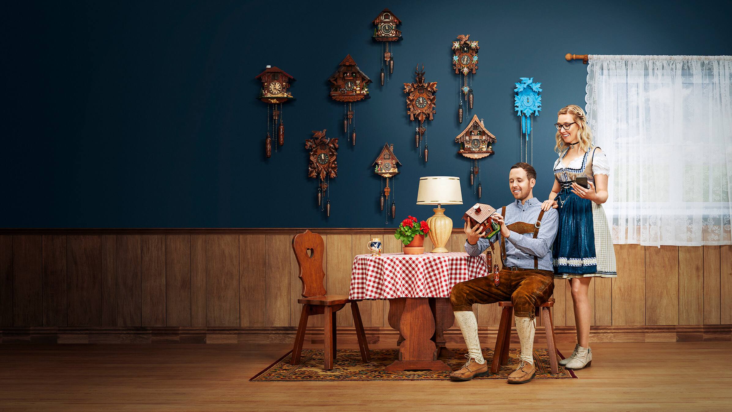 Anton and Stef of German Cuckoo Clock Nest sit at a table holding a cuckoo clock with a wall of clocks behind them.