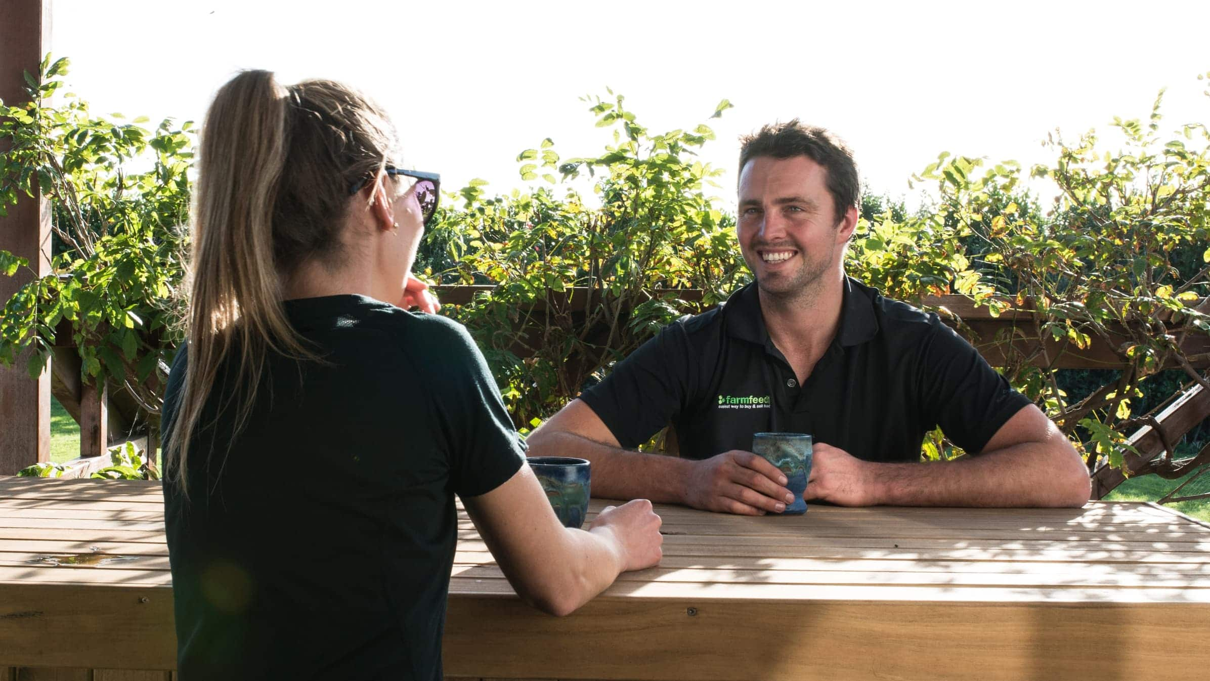 Siman Washer of Farmfeed sits with a cup at an outdoor wooden table chatting to a lady.