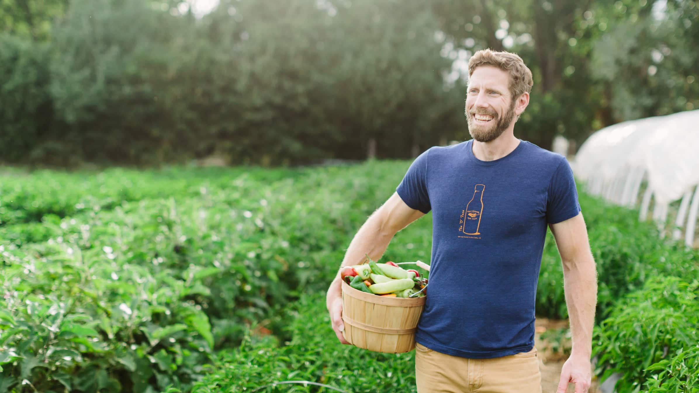 John Comeau, owner of Horsetooth Hot Sauce, standing in a plantation field holding a basket of peppers.