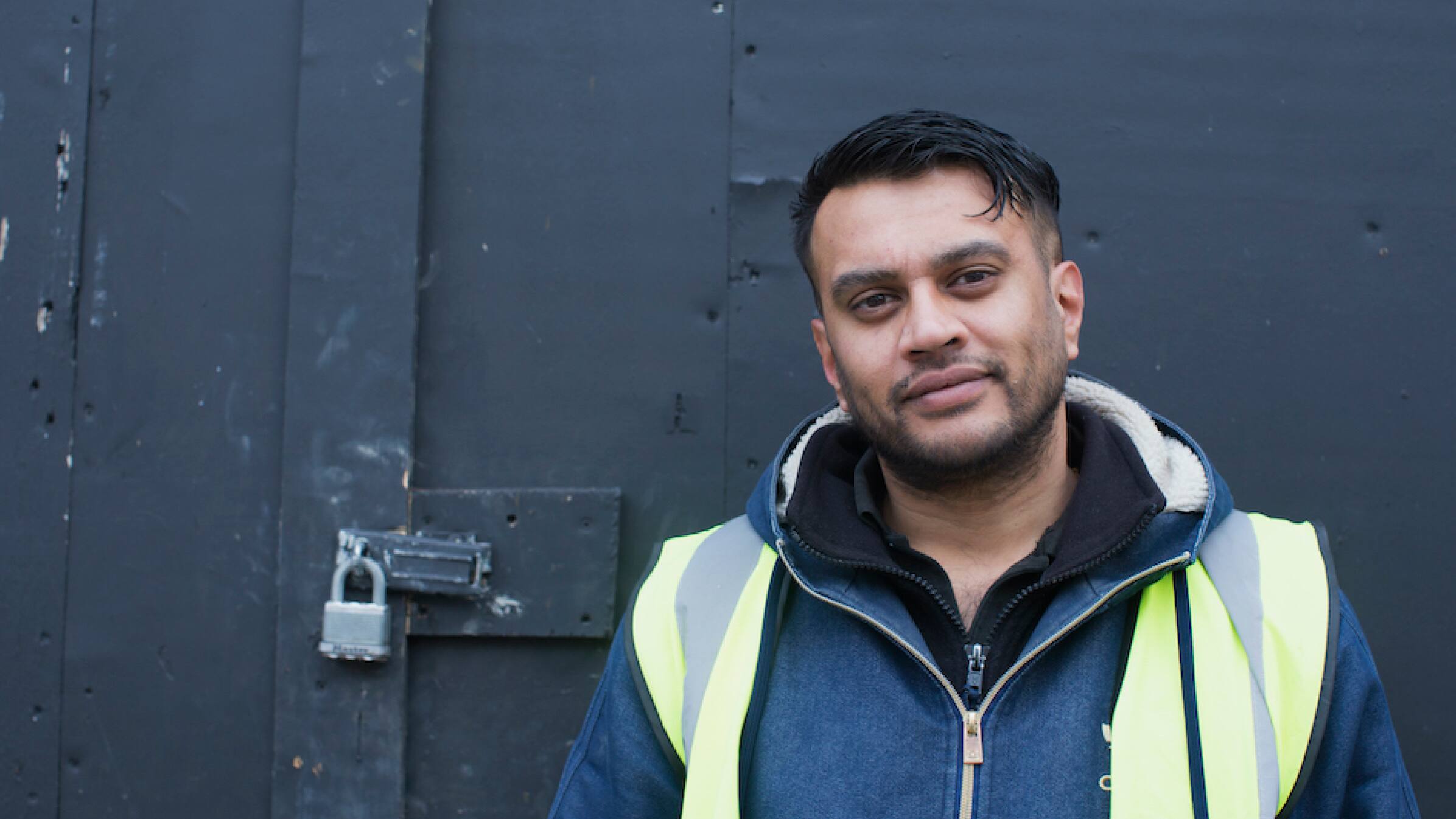Hitesh Kanbi, founder of construction company IKPE Ltd, standing in front of a warehouse door wearing a high-vis vest.