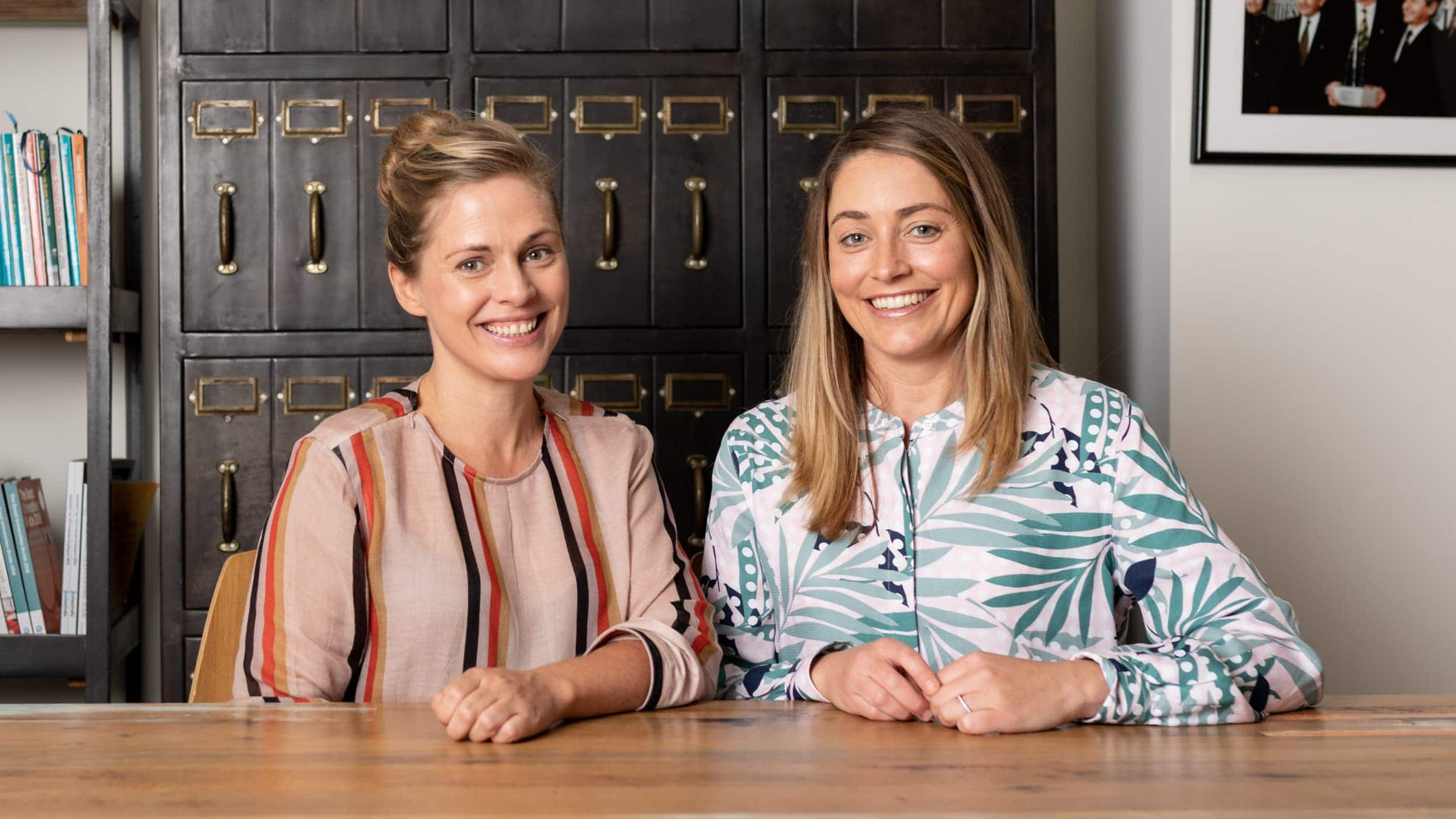 Lucy and Heidi of Mentorloop sit by each other at their office desk, smiling at the camera.