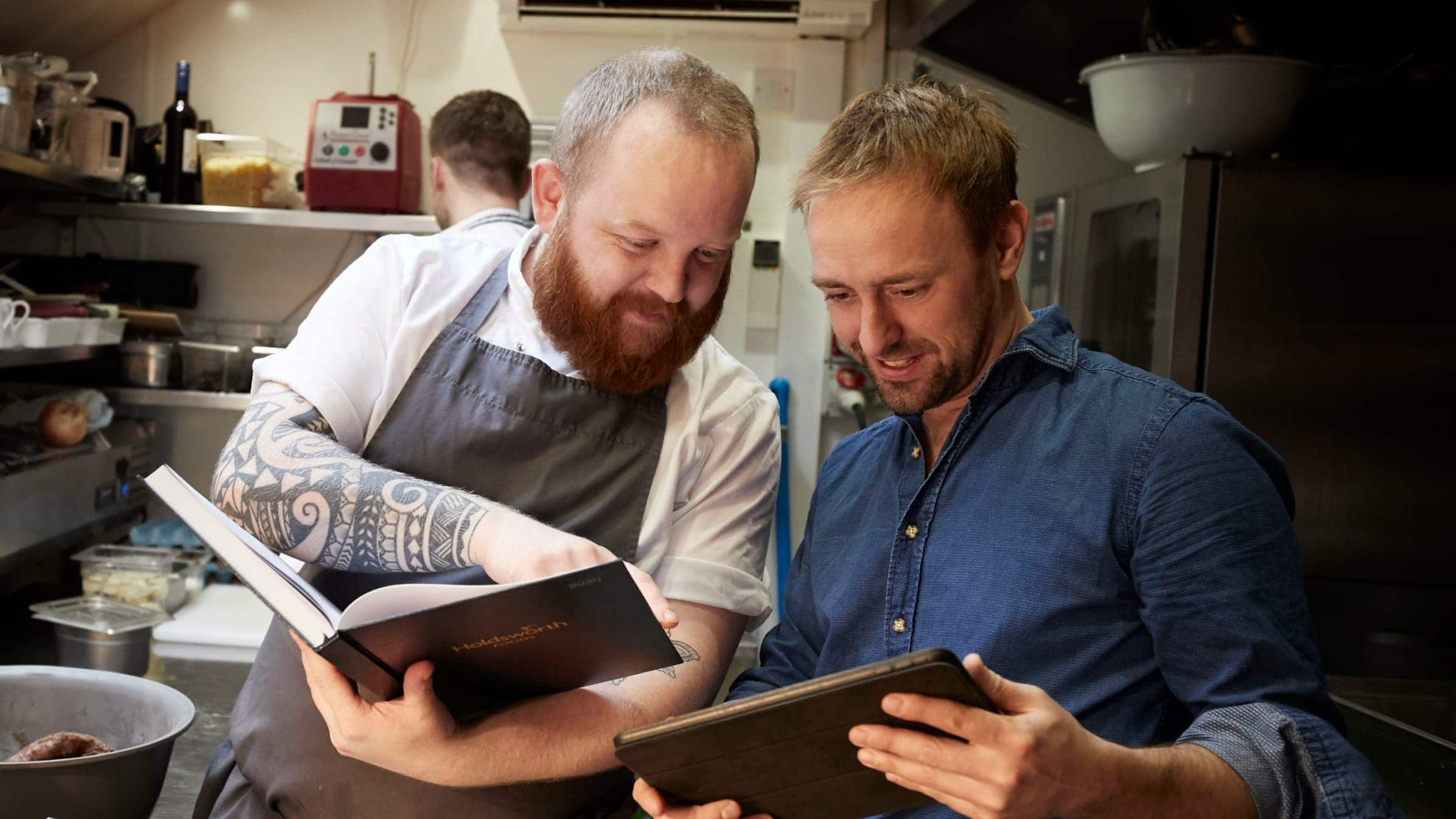 Dan Capper and colleague stand in a kitchen next to each other, looking through the pages of books they’re each holding.