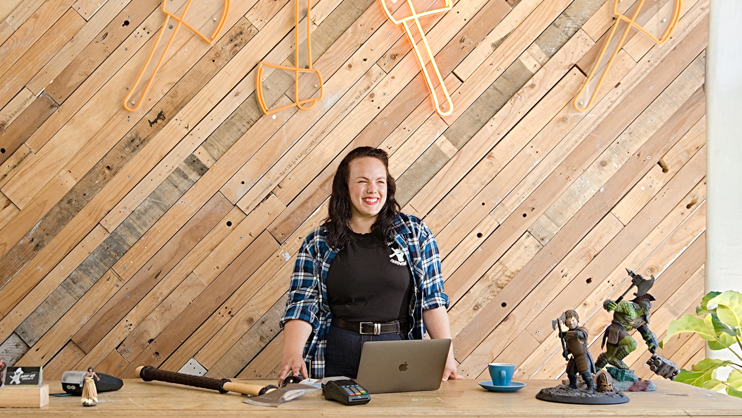 Sarah Hilyard behind a laptop in their office with an axe on the bench and axe artwork on the wall behind.