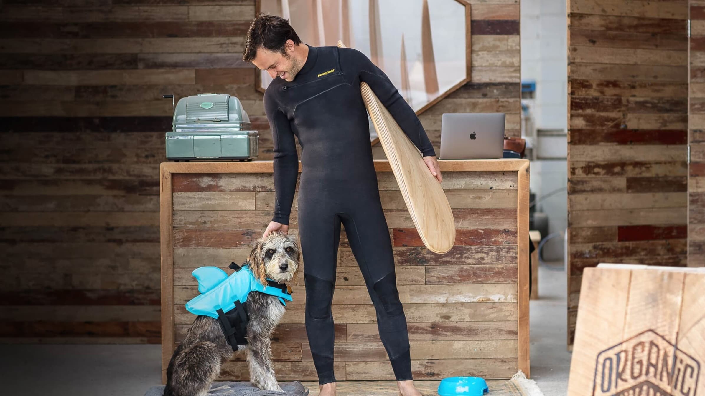 Jack Candlish, founder of Verdure Surf, wearing a wetsuit and holding a surfboard next to a dog wearing a swimming vest.