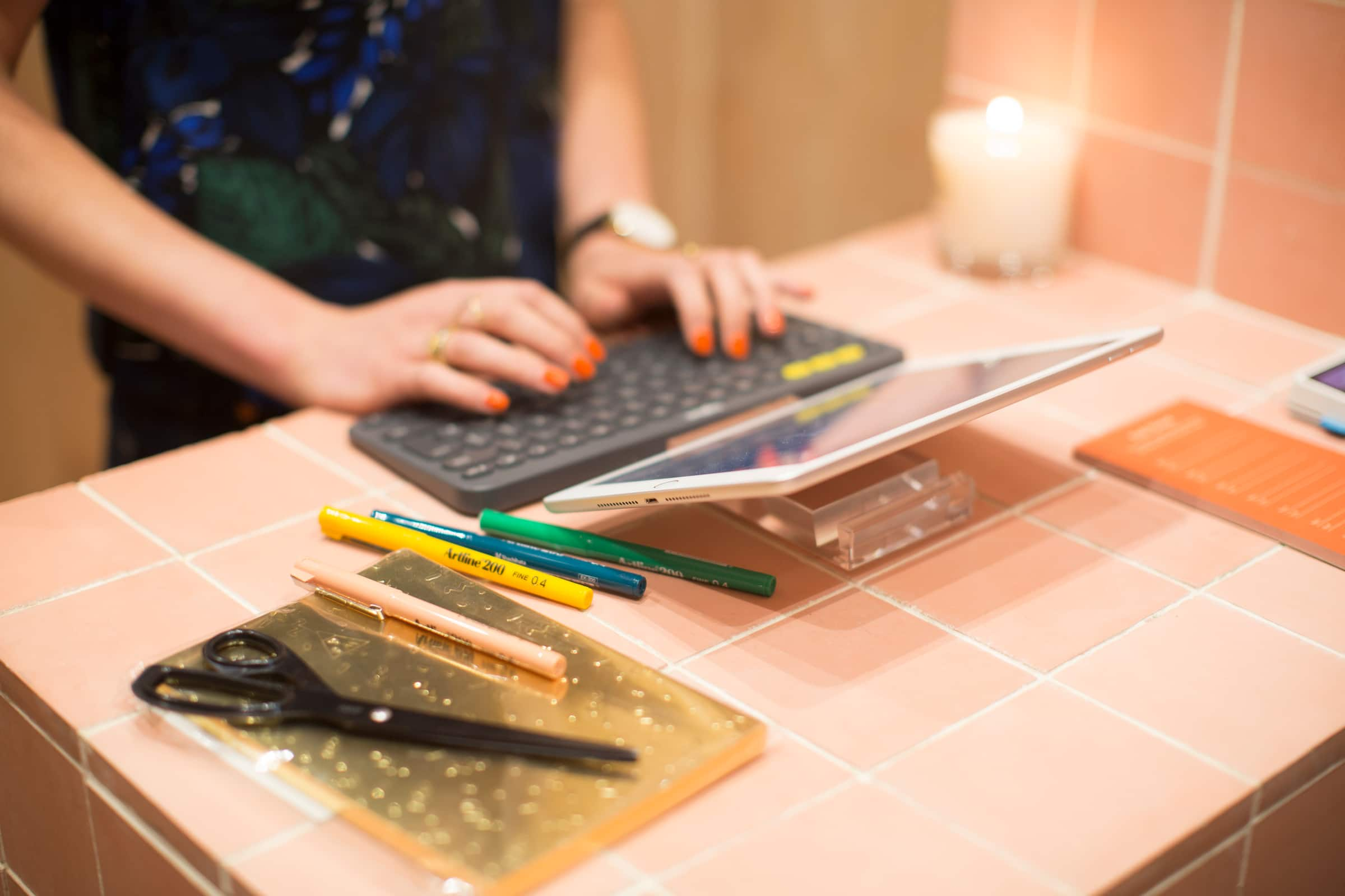 Close up on Sidonie’s hands on a keyboard with brightly painted nails and stationary pens scattered on the desk.