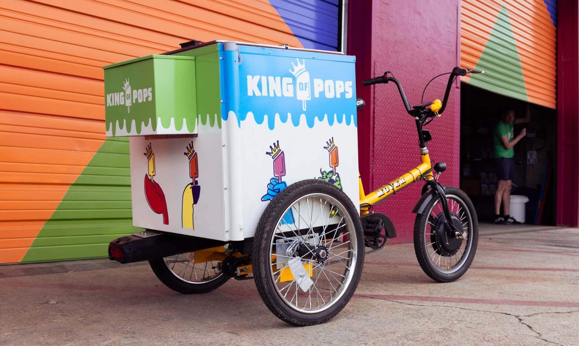A tricycle ice cream cart with "King of Pops" branding is parked in front of a colorful building with an open garage door.