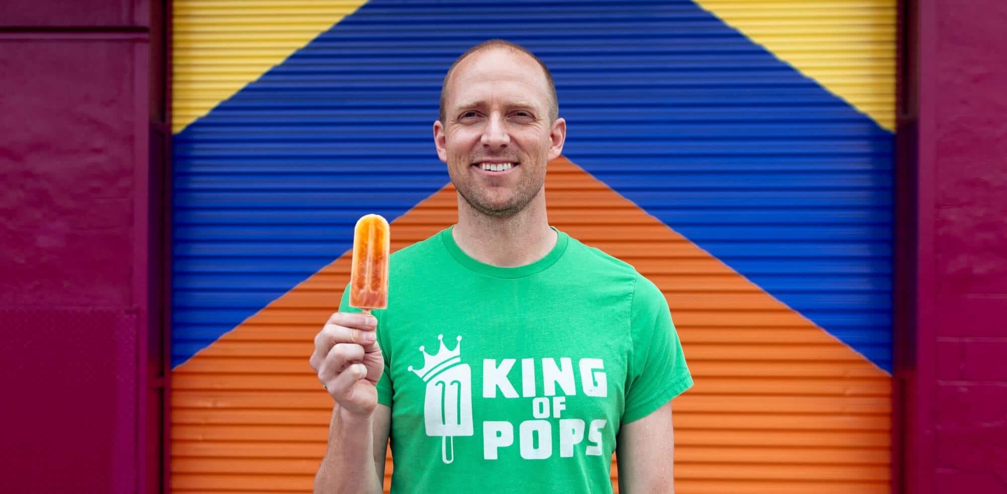 King of Pops owner Steven Carse stands in front of colorful mural with yellow, blue, and orange patterns holding a popsicle.