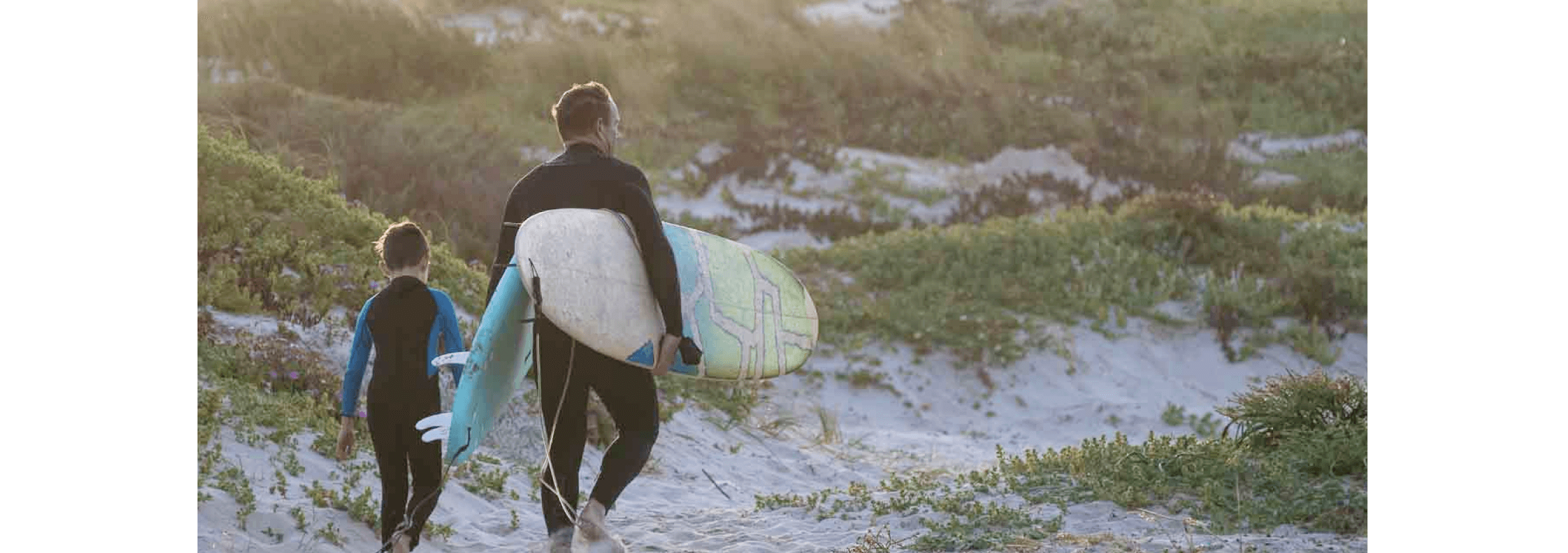 Paul Cartmel, with a surfboard under each arm. and his son walk through sand dunes in their wetsuits.