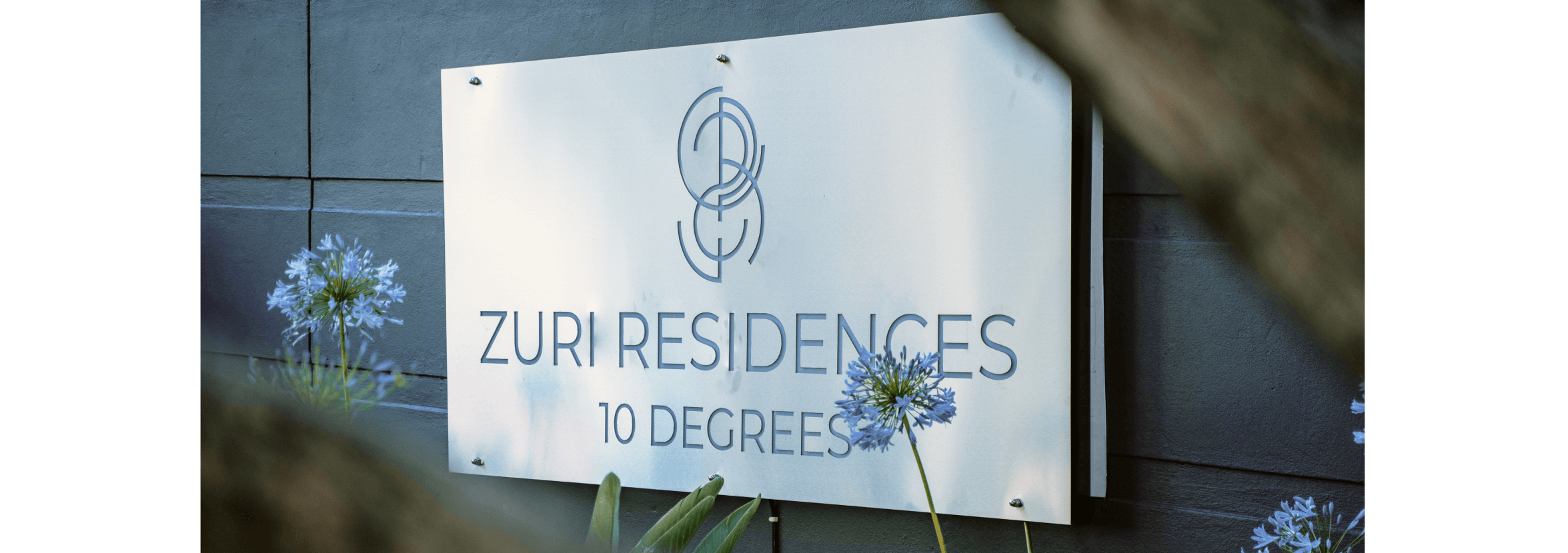 A sign with agapanthus flowers in front of it showing the words Zuri Residences, 10 degrees.
