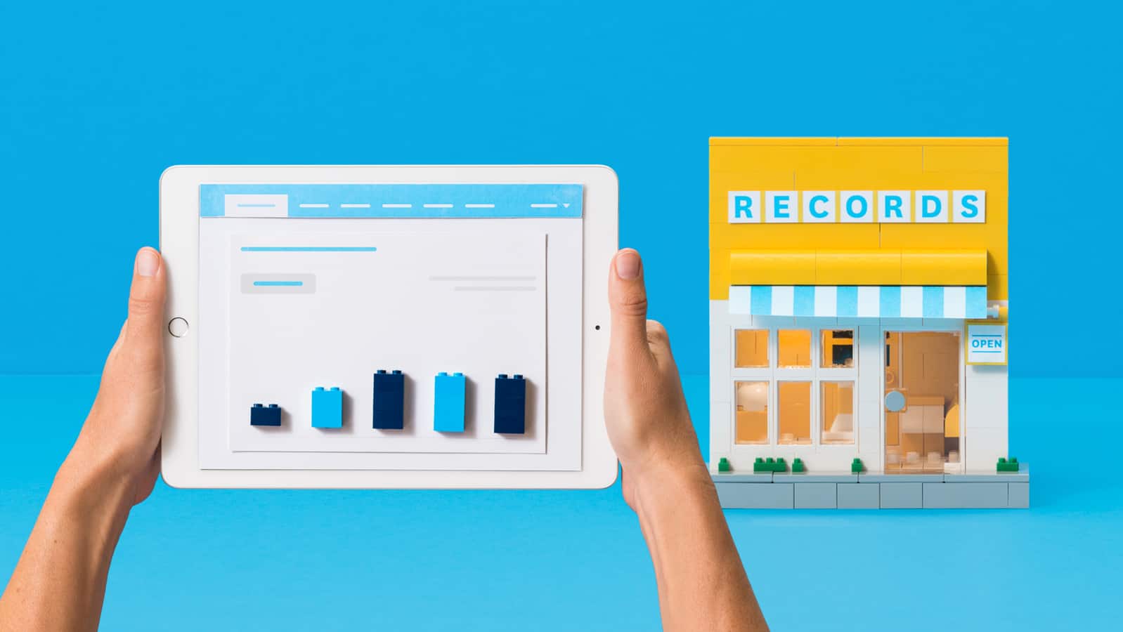 A person's hands holding a tablet, with a store made of lego in the background