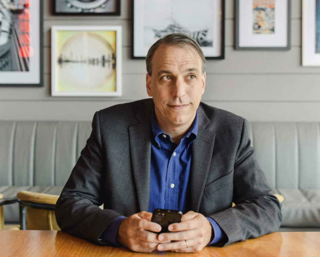 A person wearing a suit, sits at a table with a mobile phone.