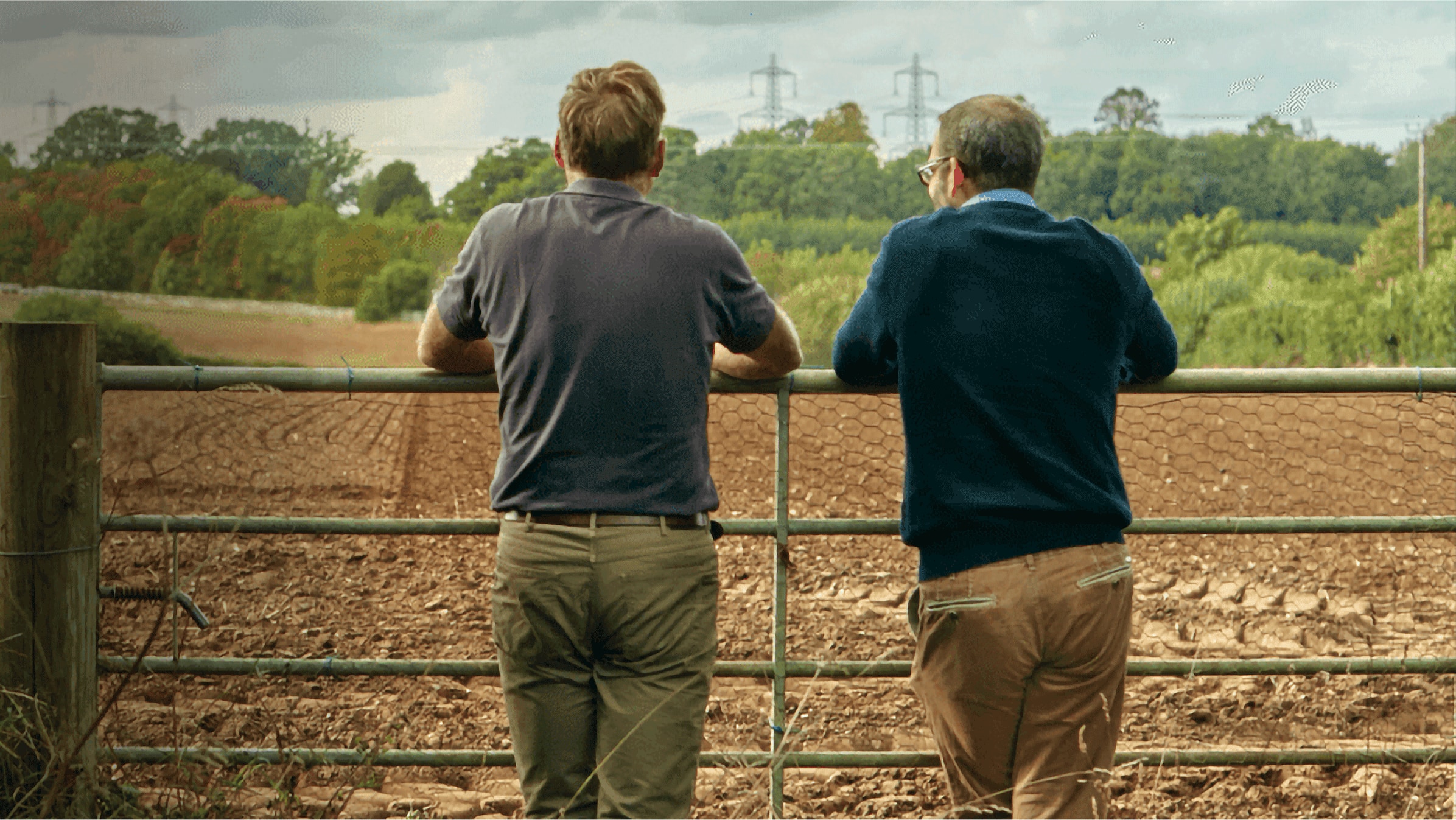 A farmer and his accountant lean on a gate overlooking a ploughed field.