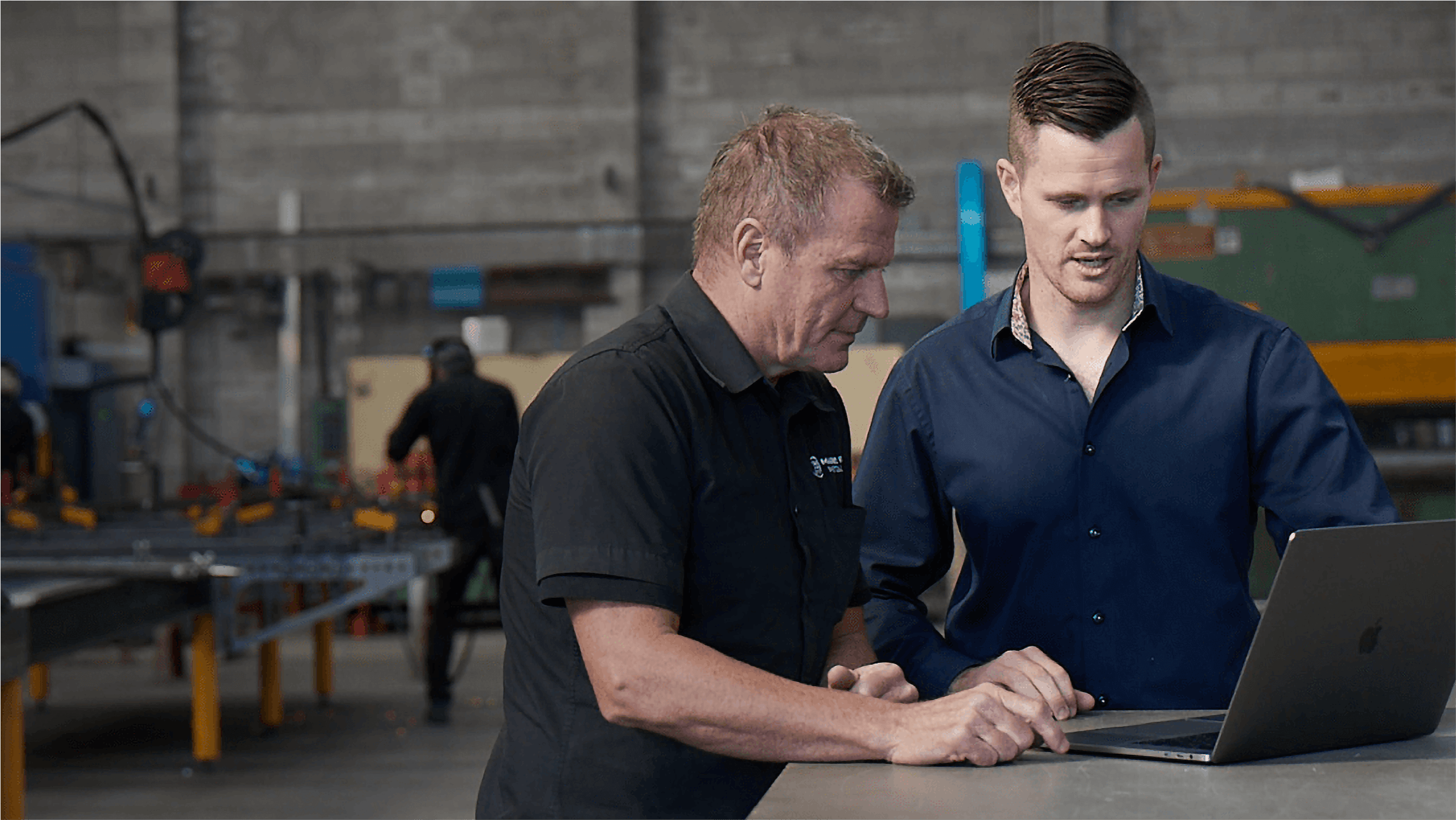 An accountant works  with a client on their laptop on site in the client’s workshop. 