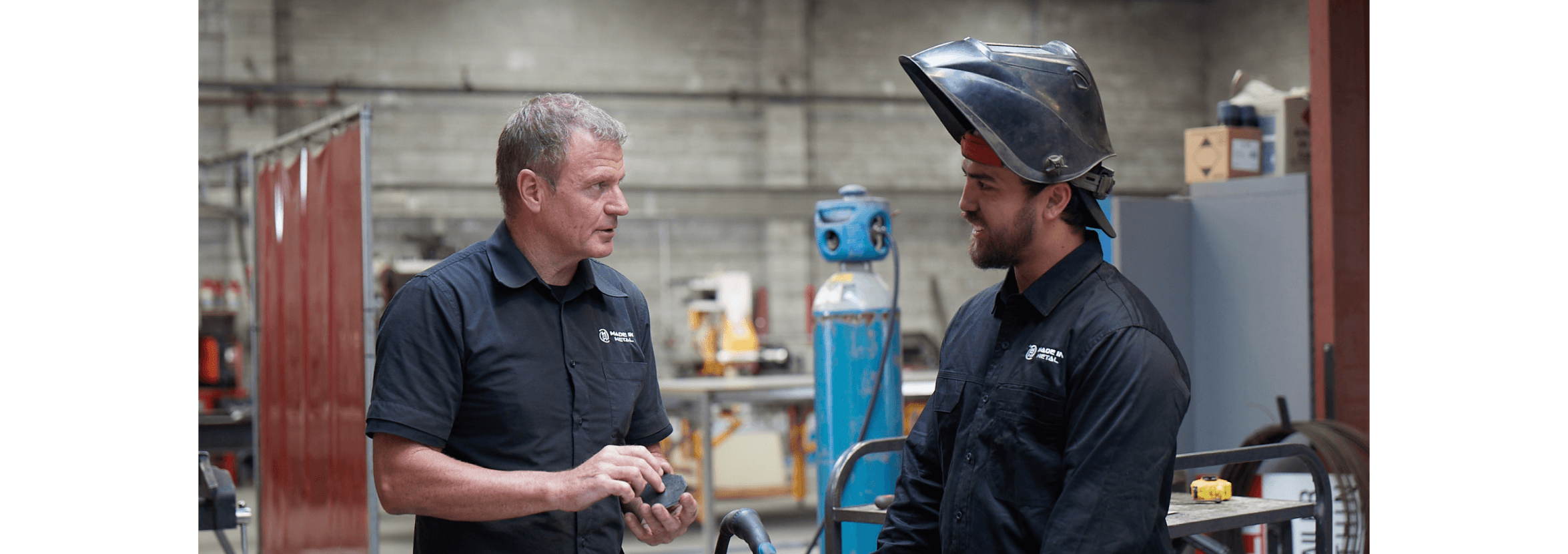 A small business owner talks with one of their staff who’s wearing a welding mask.